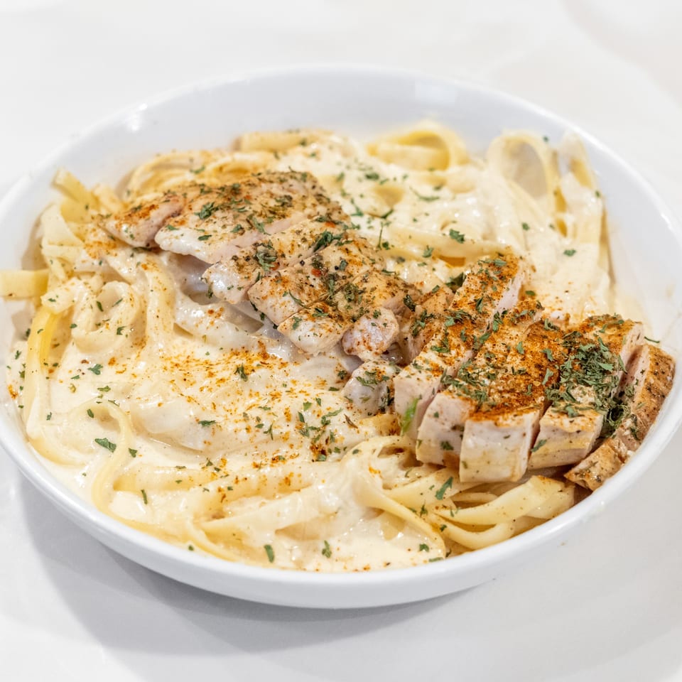 Creamy fettuccine alfredo topped with sliced chicken breast, paprika, and parsley in a white bowl.