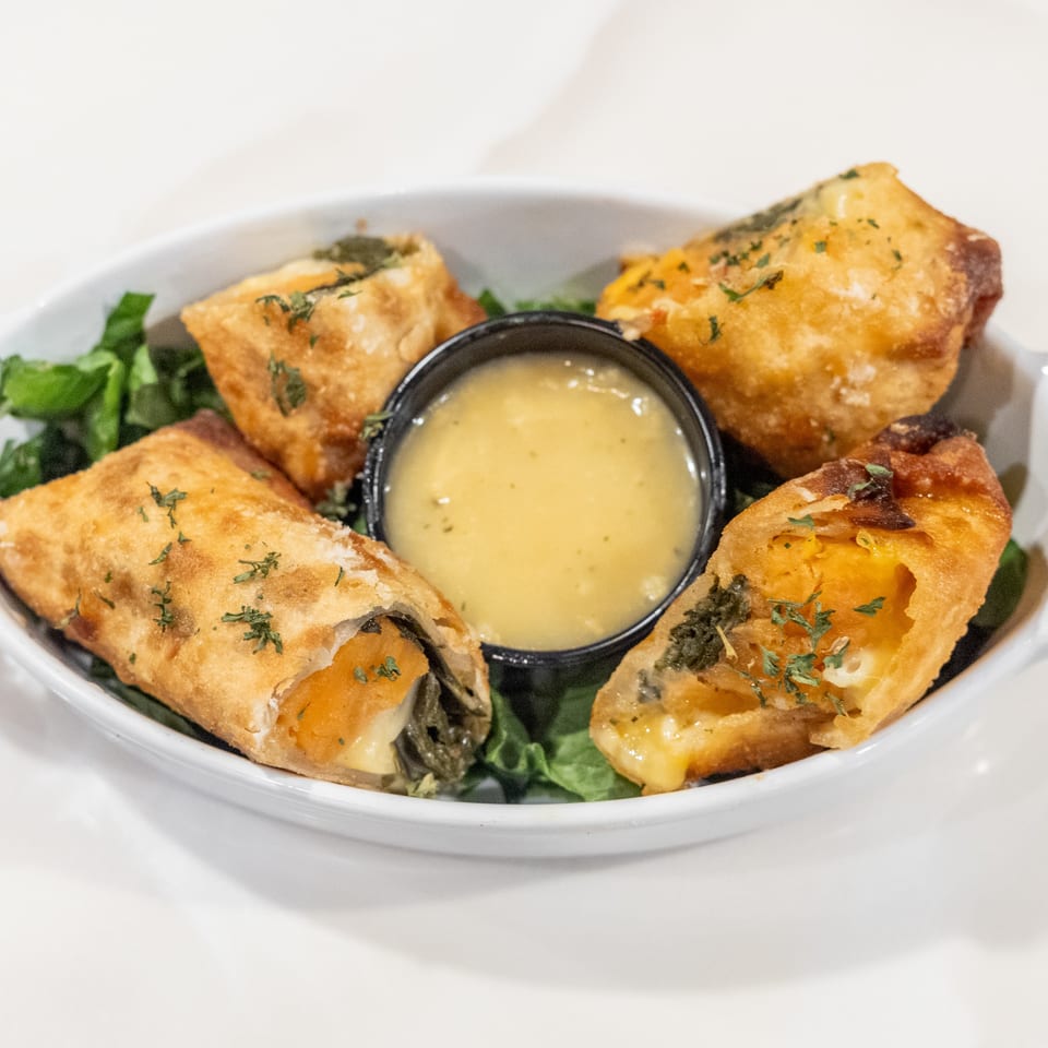 Four golden-brown egg rolls with a creamy dipping sauce, garnished with herbs, on a bed of greens.
