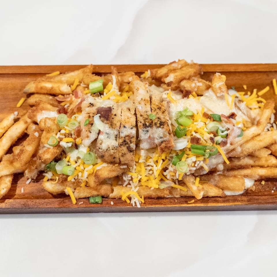 Fries topped with grilled chicken, melted cheese, bacon bits, and green onions on a wooden board.